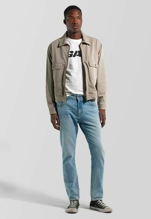 Joven con chaqueta beige, camiseta blanca con gráfico, vaqueros azul claro y zapatillas negras, posando frente a un fondo gris liso.