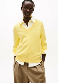 Geel V-hals trui van een zachte brei, gedragen over een witte blouse met kraag. De trui heeft een klein logo en is gecombineerd met beige geplooide broeken.