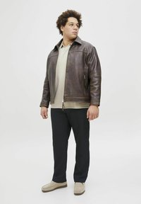 Brown leather jacket with a structured collar, worn over a beige polo shirt, paired with black trousers and beige slip-on shoes.
