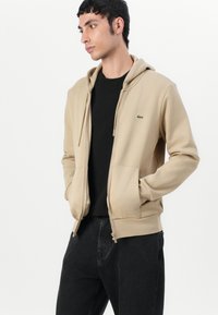 Felpa con cappuccio e zip beige, dotata di tasca a marsupio e accentuazione del logo verde. Indossata sopra una maglietta nera e abbinata a jeans scuri.