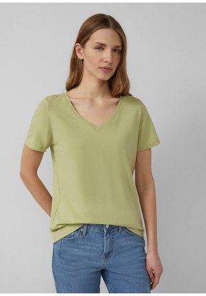 Kobieta o jasnobrązowych włosach, ubrana w jasnooliwkowy T-shirt z dekoltem w serek i niebieskie dżinsy, na tle jednolitego szarego tła.