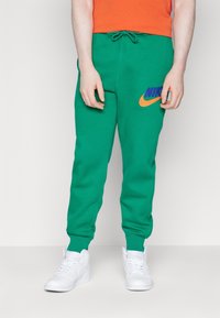 Zelené Nike tepláky s páskem na stažení, žebrovanými manžetami a vyšívaným logem ve fialové a oranžové barvě. Nošeny s bílými teniskami.