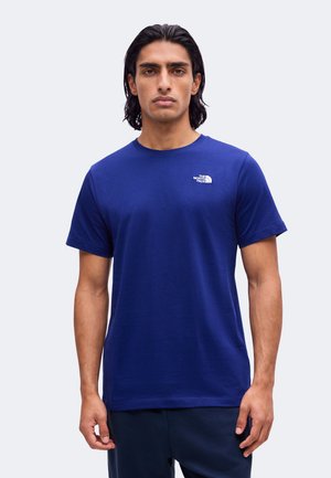 Man met halflang donker haar die een effen blauwe T-shirt met korte mouwen draagt met een klein wit North Face-logo op de borst.