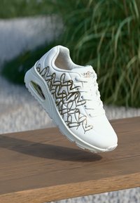 White sneaker with gold embroidered accents, mesh material, rounded toe, and cushioned sole. Features branded tongue and lace-up design.