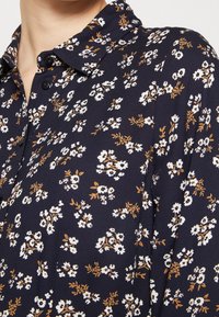 Camicia blu navy con un motivo floreale in accenti bianchi e ocra, colletto con bottoni e tessuto leggero.