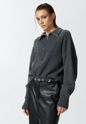 Maglione grigio a costine con colletto a mezza zip, abbinato a pantaloni in pelle nera a vita alta e una cintura nera con dettagli in metallo argentato.