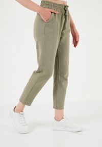 Khaki jogger hlače s elastičnim pojasom, pertlama, bočnim džepovima i suženim nogavicama, u kombinaciji s bijelim tenisicama na bijeloj pozadini.