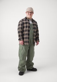 Jeune homme atteint de trisomie 21 portant des lunettes, un bonnet beige, une chemise à carreaux, une salopette verte et des chaussures noires, debout devant un fond blanc uni.