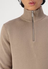 Pulover bej cu fermoar, cu guler înalt, dintr-un material tricotat texturat, cu detalii metalice argintii la fermoar. Designul ajustat evidențiază linia gâtului și umerii.