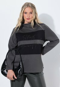 Pull color block gris et noir avec manches longues, arborant un design en texte métallique. Associé à un pantalon en cuir noir sleek et un sac à main noir.