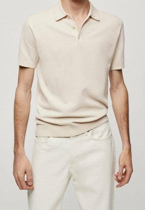 Man draagt een beige poloshirt met korte mouwen en twee knopen, gecombineerd met off-white broek, staand tegen een effen achtergrond.
