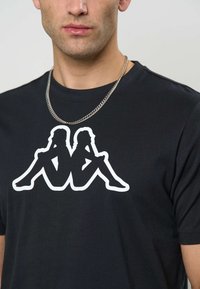 Čierne tričko s krátkym rukávom s veľkou bielou siluetou dvoch sediacich postáv na prednej strane. Viditeľný strieborný retiazkový náhrdelník.