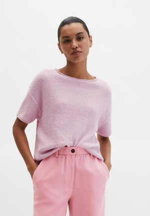 Vrouw met donker haar draagt een lichtpaars, textuurrijk T-shirt met korte mouwen, ingestopt in een hooggesneden roze broek, met haar handen in de zakken.