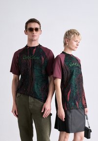 Twee individuen in bijpassende t-shirts met een donker patroon en bordeauxrode raglanmouwen, met groene tekst, gecombineerd met olijfgroene broeken en een zwarte rok.