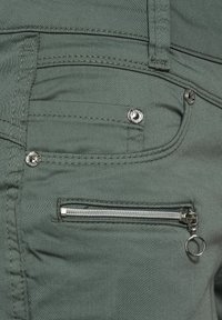 Gröna tygbyxor med en framficka med en silverknapp, en extra ficka med dragkedja och metalldetaljer, samt detaljerad sömnad.