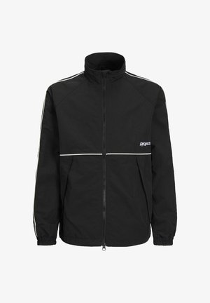 Fekete cipzáras dzseki fehér díszítéssel, fehér pólóval, szürke farmernadrággal és fekete-fehér sportcipővel párosítva. A modell egy sima háttér előtt áll.