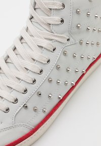 Baskets montantes en cuir blanc avec lacets crème, clous métalliques argentés sur le côté et bande en daim rouge au-dessus de la semelle.