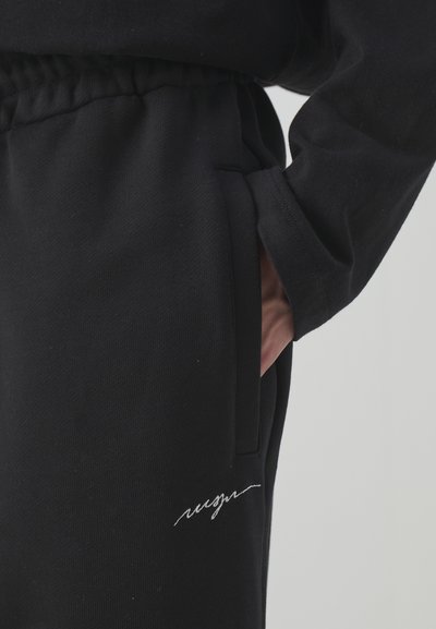 Sorte sweatpants med ribbet talje, sidelommer og hvid broderet tekst. Stoffet fremstår blødt med en struktureret overflade.