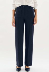Femme portant un pantalon droit bleu marine avec des poches plaquées à l'avant, associé à des ballerines noires à bout pointu et une chemise blanche à manches longues.