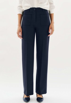 Femme portant un pantalon droit bleu marine avec des poches plaquées à l'avant, associé à des ballerines noires à bout pointu et une chemise blanche à manches longues.