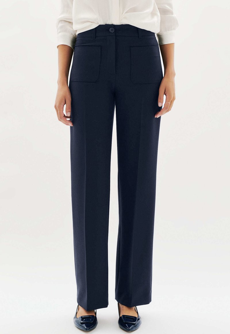 Femme portant un pantalon droit bleu marine avec des poches plaquées à l'avant, associé à des ballerines noires à bout pointu et une chemise blanche à manches longues.