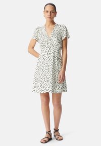 Abito bianco con pois neri, maniche a sbuffo corte, scollo a V, davanti con bottoni, gonna svasata. Indossato con sandali neri con le fasce.