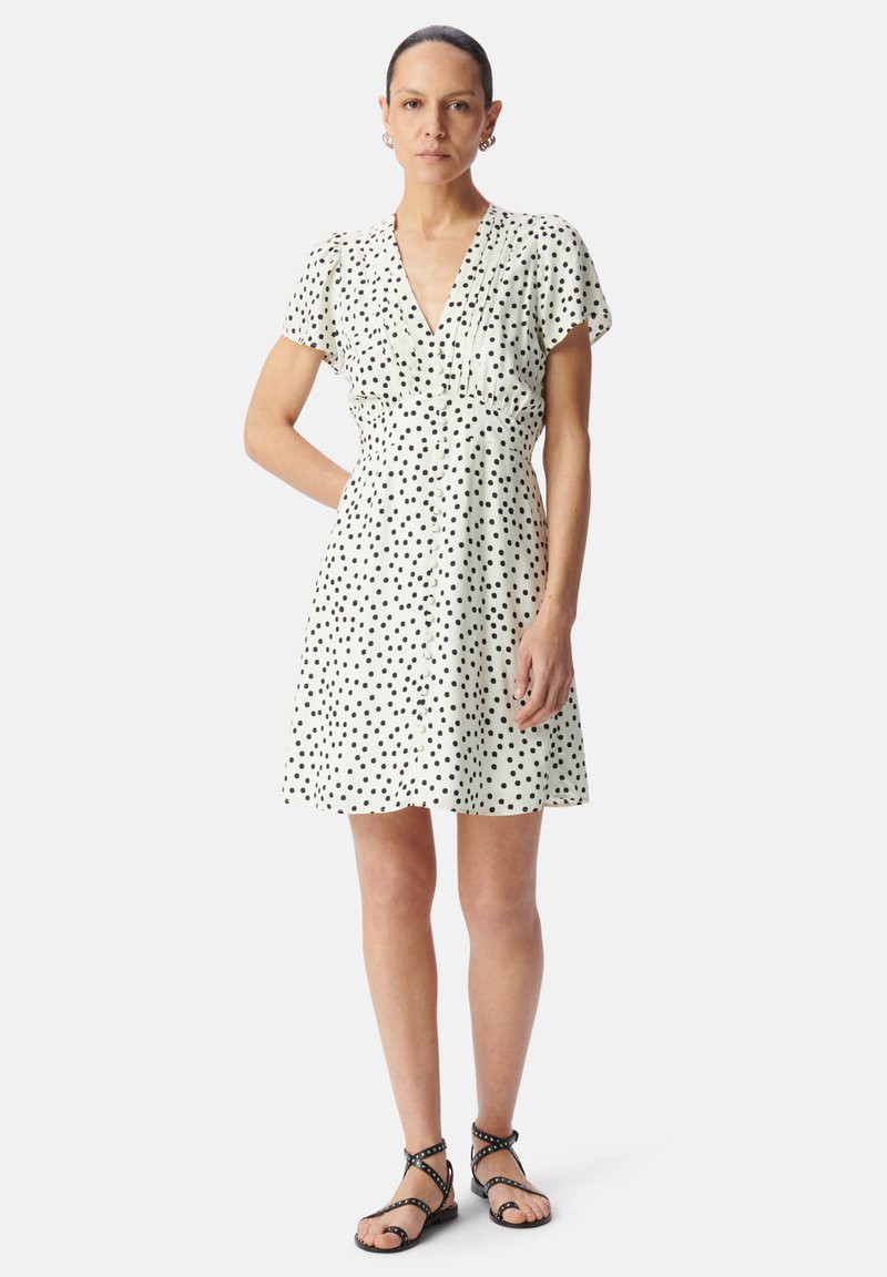 Witte jurk met zwarte stippen, korte pofmouwen, V-hals, knoopsluiting aan de voorkant, uitlopende rok. Draagt met zwart strap-sandalen.