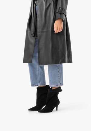 Personne portant un jean bleu raccourci, des bottines noires en daim à talon et un trench-coat en cuir noir avec ceinture et détail boucle.