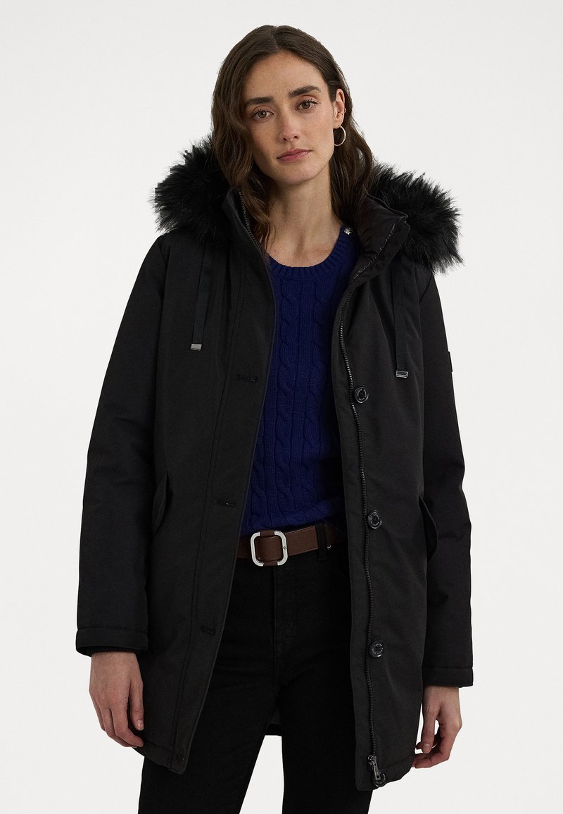Parka noire avec une capuche doublée en fausse fourrure, boutons à l'avant, fermeture éclair et poches latérales, portée sur un pull en tricot torsadé bleu marine.