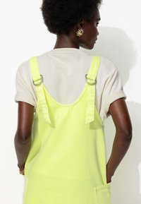 Salopette jaune fluo avec bretelles ajustables et accessoires en argent, portée par-dessus une chemise à manches retroussées beige clair. Détail de poche simple.