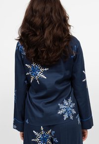 Granatowa bluzka z długim rękawem z haftem w kształcie płatków śniegu w niebieskim i białym kolorze, o gładkiej fakturze oraz kontrastowych jasnoniebieskich akcentach.