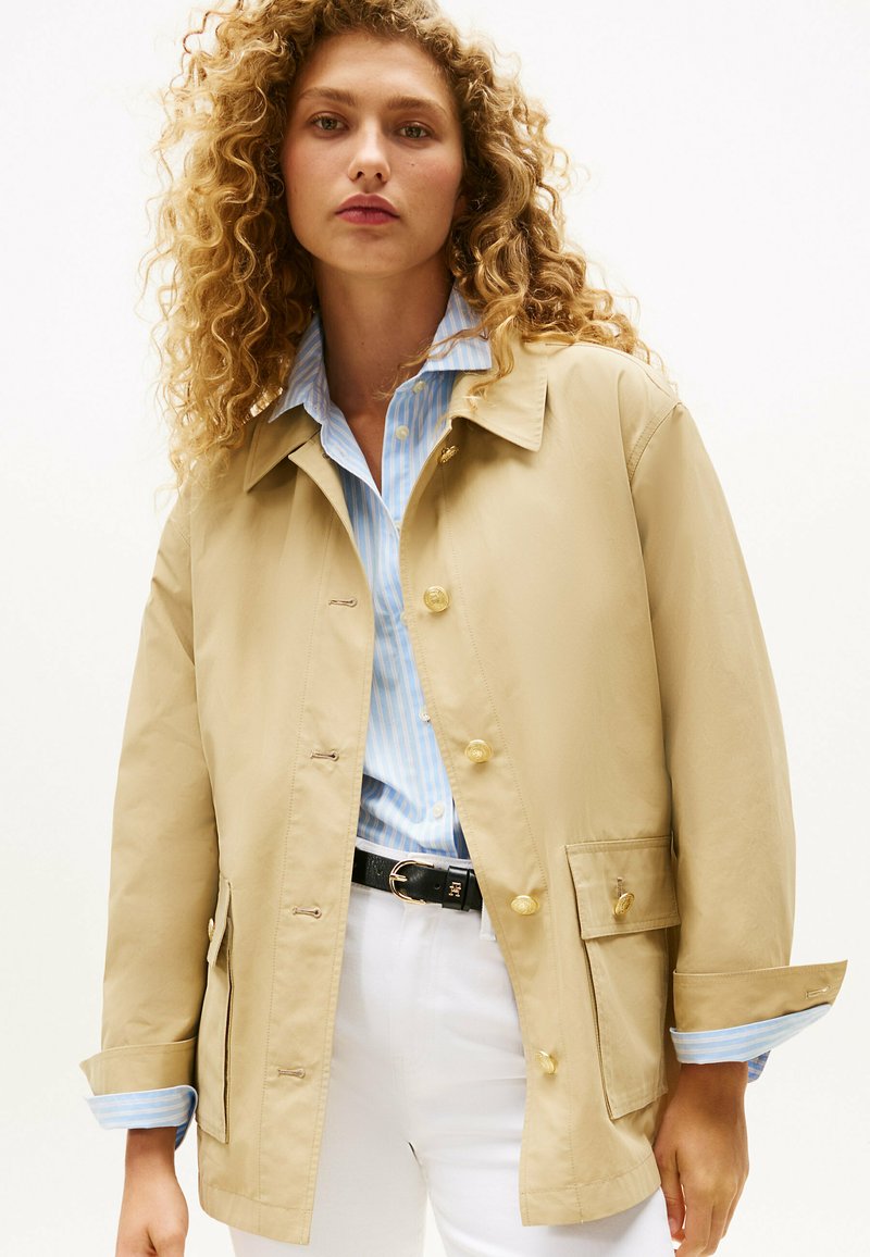 Beige waterdichte jas met een kraag, gouden knoopsluiting, twee zijzakken en een losse pasvorm. Draagt over een blauw gestreept shirt.