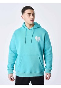 Sudadera turquesa con bolsillo frontal, capucha con cordón y un gráfico de un corazón roto en el pecho. Tejido suave con un ajuste holgado.