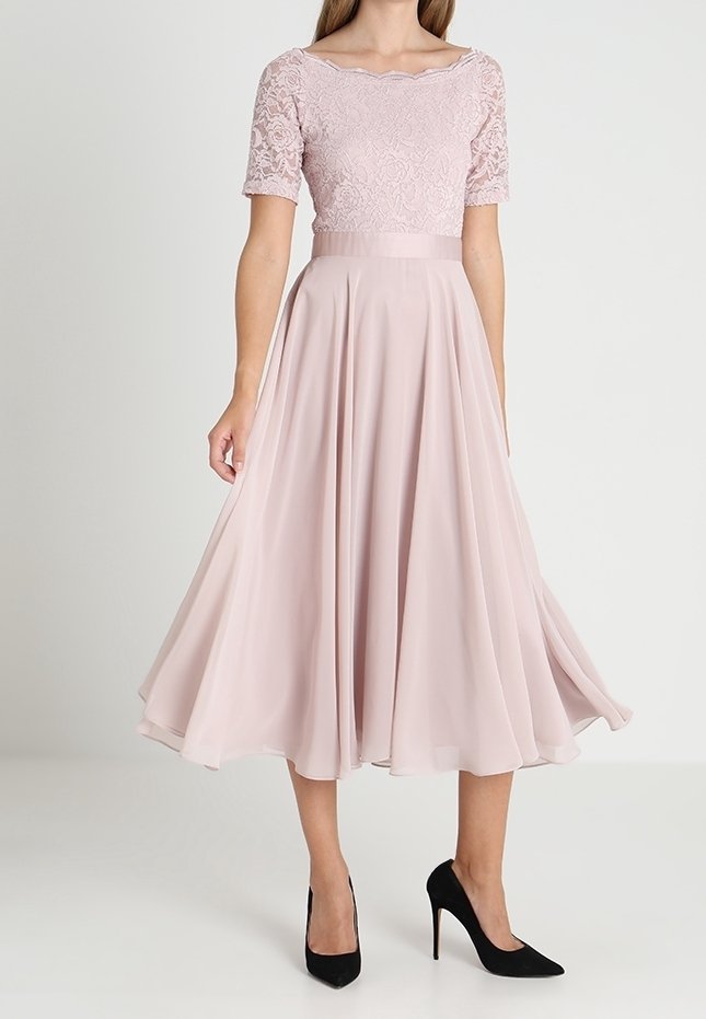 Femme portant une robe à top en dentelle rose pâle avec une jupe midi fluide et des escarpins noirs, debout devant un fond uni.