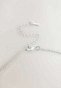 Zilveren kettingarmband met ronde schakels, een kreeftsluiting en een klein label eraan bevestigd, afgezet tegen een lichte achtergrond.
