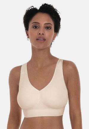 Frau mit kurzen, lockigen Haaren, die einen nahtlosen, hellbeigen BH mit breiten Trägern vor einem einfarbigen Hintergrund trägt.