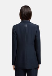 Blazer sartorial blu navy con vestibilità slim, revers a lancia e etichetta di marca sul retro. Texture liscia e design strutturato.