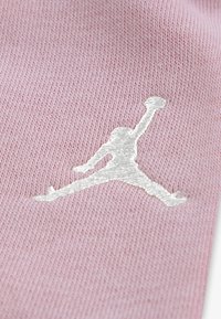 Jasnoróżowy materiał tekstylny z białym haftowanym logo Jumpman, prezentujący miękką dzianinę i gładką fakturę.