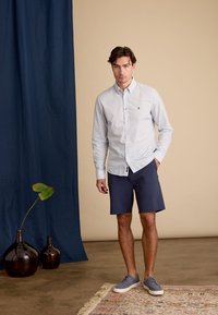 Licht gepatternde knoopshirt in lichtblauw, gecombineerd met marineblauwe shorts. Model draagt grijze schoenen; de achtergrond heeft een donkerblauwe gordijn en een decoratief tapijt.