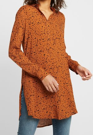 Vrouw draagt een oranje tuniekblouse met zwarte vlekken en lange mouwen en een blauwe spijkerbroek, staand tegen een effen achtergrond.
