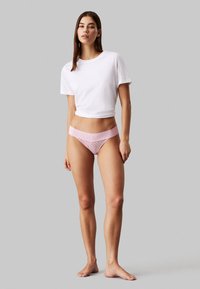 Tanga de renda rosa com um cinto largo e decorativo, combinada com uma t-shirt cropped branca simples. Tecido suave, design minimalista.