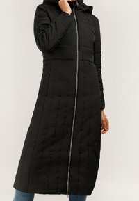 Czarny długi pikowany płaszcz z kapturem i srebrnym zamkiem z przodu, noszony na niebieskich dżinsach, z jedną ręką przy twarzy, a drugą swobodnie opuszczoną.