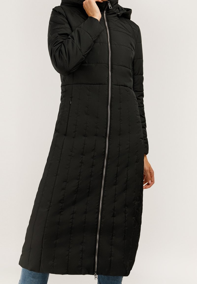 Czarny długi pikowany płaszcz z kapturem i srebrnym zamkiem z przodu, noszony na niebieskich dżinsach, z jedną ręką przy twarzy, a drugą swobodnie opuszczoną.