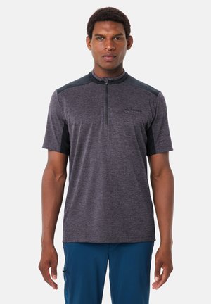 Homme portant un maillot de sport gris et noir à manches courtes avec fermeture éclair, associé à un pantalon bleu, debout devant un fond clair uni.