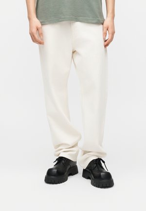 Mand iført løstsiddende hvide bukser og sorte chunky snøresko, stående med afslappede hænder ned langs siderne mod en ensfarvet baggrund.