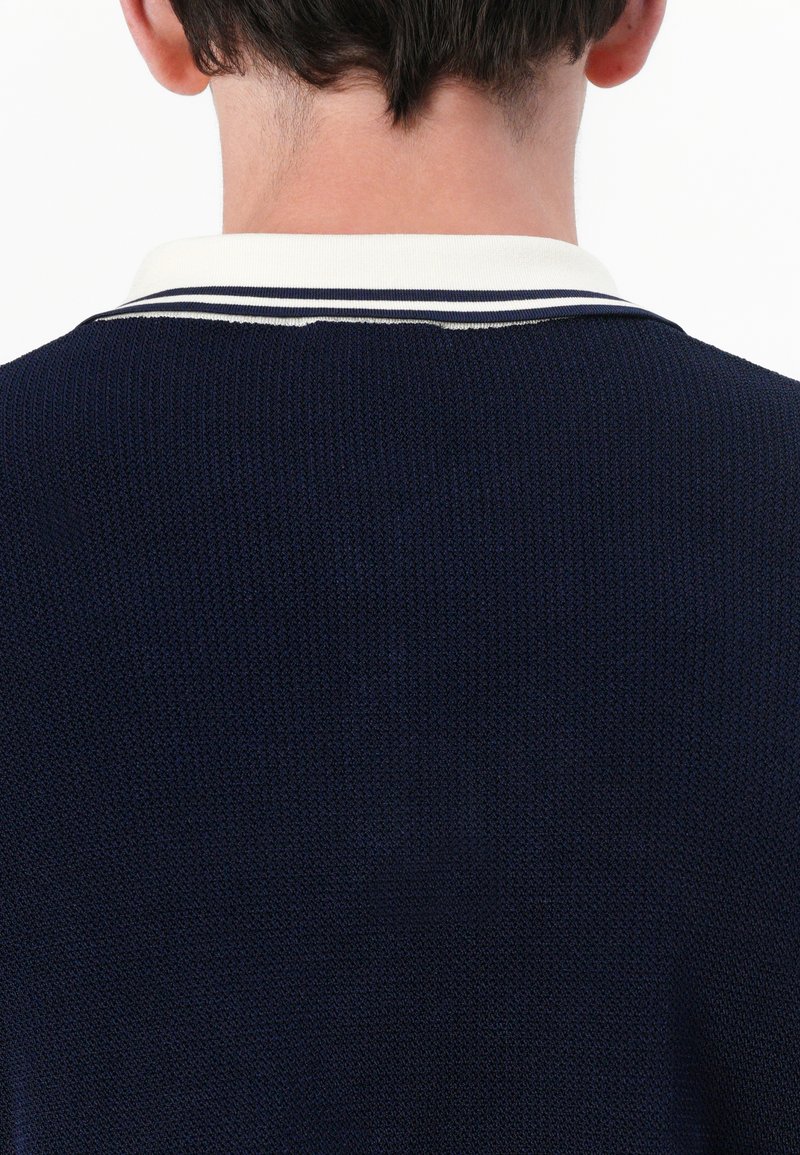 Navy-blaues Strick-Polohemd mit weißem Kragen, das zwei dunkelblaue Streifen aufweist. Der Stoff wirkt strukturiert und figurbetont am Kragen.