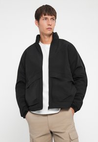 Black bomber jacket with a high collar, front zip closure, and two front pockets, made from smooth, lightweight fabric.