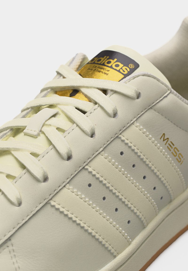 Gros plan d'une basket Adidas couleur crème avec logo doré sur la languette, les trois bandes emblématiques, et "MESSI" inscrit en lettres dorées sur le côté.