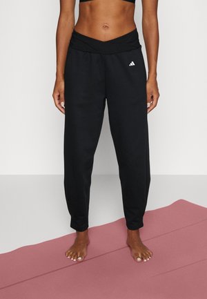 Woman wearing black Adidas joggers standing barefoot on a pink yoga mat against a plain background.