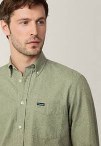 Camisa de botones verde claro hecha de una tela texturizada. Presenta un bolsillo en el pecho y un pequeño parche de marca cerca del bolsillo.